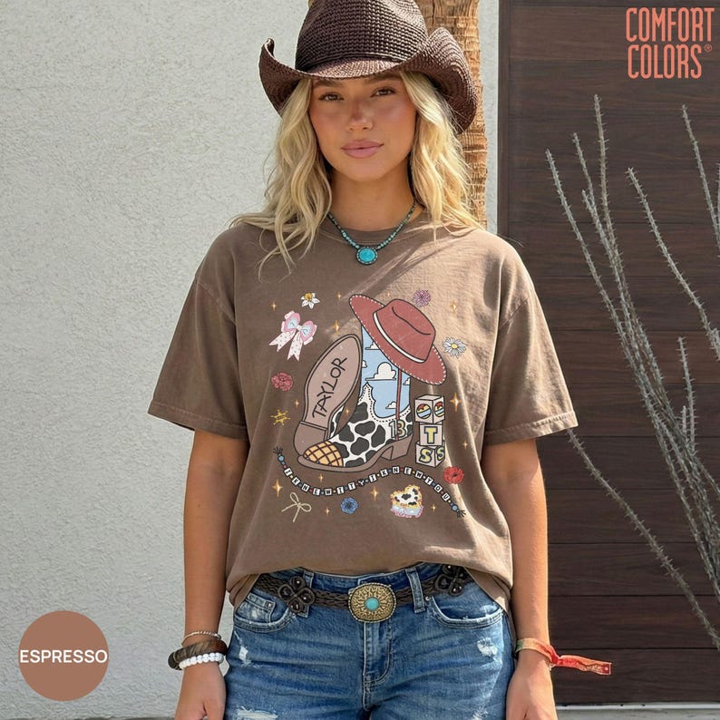 May include: A brown short-sleeve t-shirt featuring a graphic print of a cowboy boot, a cowboy hat, a bow, and the text "TAYLOR" and "13". The shirt is paired with blue denim jeans and a brown belt with a large decorative buckle.