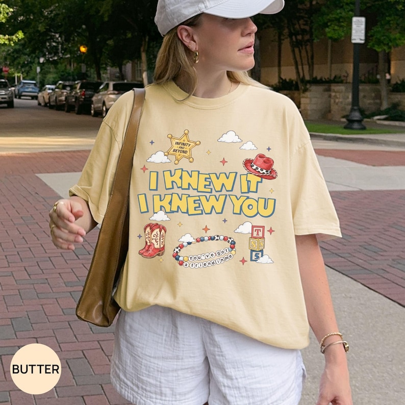 Op de afbeelding: Een boterkleurig t-shirt met een grafische print met de tekst "I KNEW IT I KNEW YOU". Het ontwerp bevat een sheriffster, een cowboyhoed, rode laarzen, een kralenarmband met de tekst "YOU'VE GOT A FRIEND IN ME" en speelgoedblokken.