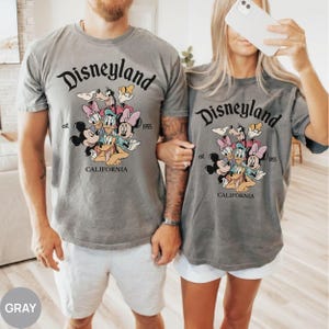 以下が含まれることがあります： ディズニーランドのロゴとディズニーキャラクターのグラフィックが特徴のグレーのTシャツ。「Disneyland」の文字がキャラクターの上にアーチ状に配置され、その下に「est. 1955」と「CALIFORNIA」と書かれています。半袖のシャツです。