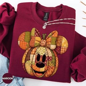 Op de afbeelding: Een bordeauxrood sweatshirt met lange mouwen en een grote afbeelding van een pompoen in de vorm van een muizenkop met een strik. De pompoen is samengesteld uit diverse herfstkleurige patchwork-stofpatronen in oranje, geel, bruin en groen.