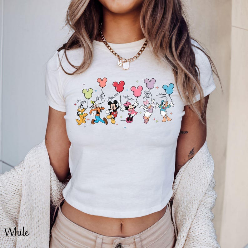 May include: White crop top featuring a colorful graphic of Disney characters holding balloons. The characters include Pluto, Goofy, Mickey Mouse, Minnie Mouse, Daisy Duck, and Donald Duck. Each character is holding a balloon with their signature color.