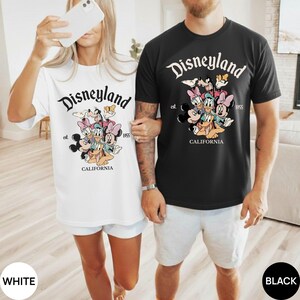 以下が含まれることがあります： ディズニーランドのグラフィックが特徴の白と黒のTシャツ。デザインには、ディズニーランドのロゴ、さまざまな漫画のキャラクター、「CALIFORNIA」と「1955」の文字が含まれています。白いシャツは白いショートパンツと、黒いシャツはグレーのショートパンツと組み合わせています。