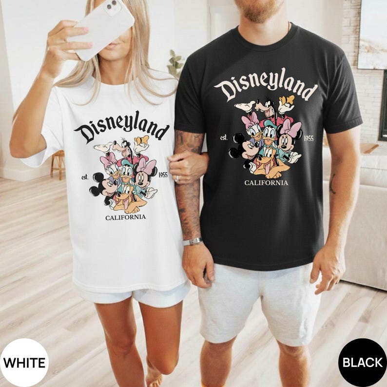 以下が含まれることがあります： ディズニーランドのグラフィックが特徴の白と黒のTシャツ。デザインには、ディズニーランドのロゴ、さまざまな漫画のキャラクター、「CALIFORNIA」と「1955」の文字が含まれています。白いシャツは白いショートパンツと、黒いシャツはグレーのショートパンツと組み合わせています。