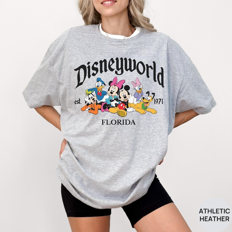 May include: An athletic heather gray t-shirt featuring a black graphic that reads Disneyworld est. 1971 FLORIDA. The design includes colorful illustrations of Mickey Mouse, Minnie Mouse, Donald Duck, Daisy Duck, Goofy, and Pluto in a group.