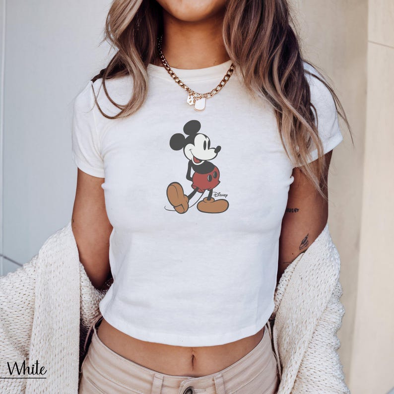 K&ouml;nnte beinhalten: Wei&szlig;es Crop-Top mit Mickey Mouse-Grafik. Die Zeichentrickfigur tr&auml;gt rote Shorts, braune Schuhe und schwarze Ohren. Das Wort "Disney" ist auf dem Shirt aufgedruckt. Das Wort "White" ist unten links aufgedruckt.