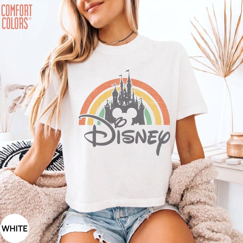 May include: A white short-sleeve t-shirt featuring a graphic print of a grey castle silhouette centered within a multi-colored rainbow arch. The word Disney is printed in a grey script font below the castle, with a Mickey Mouse icon integrated.