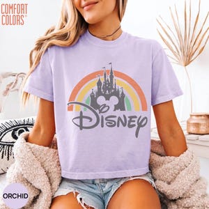 May include: A light purple short-sleeve t-shirt featuring a graphic of a castle silhouette centered in front of a multi-colored rainbow arch. The word Disney is printed in a dark gray script font below the castle. The text Comfort Colors is visible.