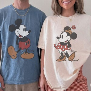 May include: Two short-sleeved t-shirts, one blue and one ivory, featuring Mickey and Minnie Mouse. The Mickey shirt is blue with a large graphic of Mickey Mouse. The Minnie shirt is ivory with a Minnie Mouse graphic. Both shirts have the Disney logo.