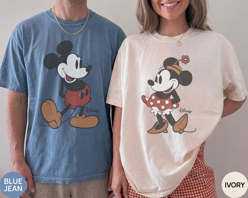 May include: Two short-sleeved t-shirts, one blue and one ivory, featuring Mickey and Minnie Mouse. The Mickey shirt is blue with a large graphic of Mickey Mouse. The Minnie shirt is ivory with a Minnie Mouse graphic. Both shirts have the Disney logo.