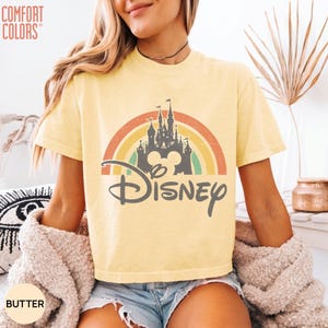May include: A butter yellow short-sleeve t-shirt featuring a graphic of a dark gray castle silhouette centered in front of a muted rainbow arch. The word Disney is printed in a dark gray script font below the castle. The text Comfort Colors is visible.