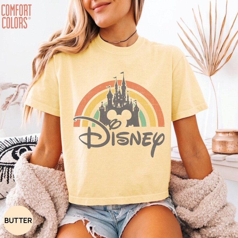 May include: A butter yellow short-sleeve t-shirt featuring a graphic of a dark gray castle silhouette centered in front of a muted rainbow arch. The word Disney is printed in a dark gray script font below the castle. The text Comfort Colors is visible.