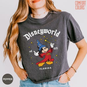 May include: Dark gray t-shirt with a graphic of Mickey Mouse in a wizard outfit, holding a wand. The shirt reads "Disneyworld est. 1971 FLORIDA" in white lettering. The shirt is a Comfort Colors brand.