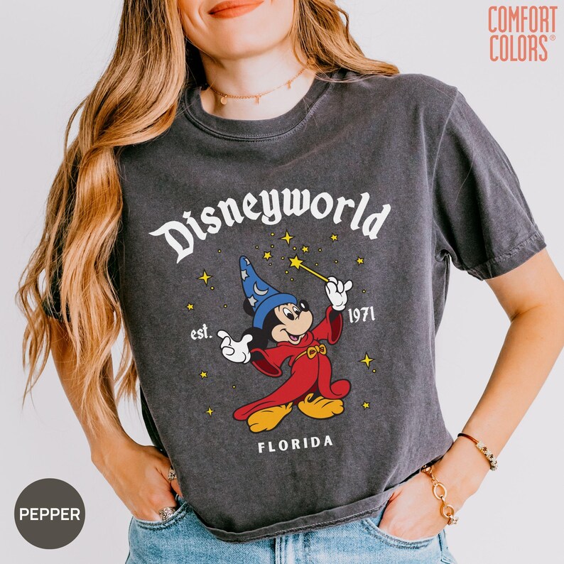 May include: Dark gray t-shirt with a graphic of Mickey Mouse in a wizard outfit, holding a wand. The shirt reads "Disneyworld est. 1971 FLORIDA" in white lettering. The shirt is a Comfort Colors brand.
