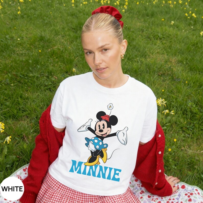 May include: A white short-sleeved t-shirt featuring a graphic of Minnie Mouse wearing a blue polka-dot skirt and yellow shoes, with the name MINNIE printed in large blue letters underneath the character illustration on the front of the garment.