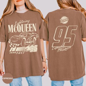 Pu&ograve; includere: T-shirt color espresso con grafica di Saetta McQueen dal film Cars. Il design frontale include il testo "Lightning McQueen", un'illustrazione di auto, il numero "95" e "Think Fast". Il retro mostra "95" e il testo "Lightning McQueen".