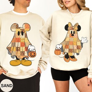 May include: Two sand-colored crewneck sweatshirts featuring graphic prints of cartoon characters dressed as ghosts. The ghosts are covered in a patchwork pattern of orange, brown, and yellow autumn-themed fabrics and are holding small orange jack-o'-lantern buckets.