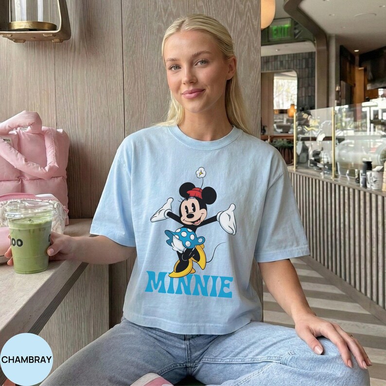 May include: A light blue short-sleeve t-shirt featuring a graphic of Minnie Mouse wearing a red hat and a blue polka-dot skirt. The character is positioned above the word MINNIE printed in bold blue capital letters on the front of the shirt.