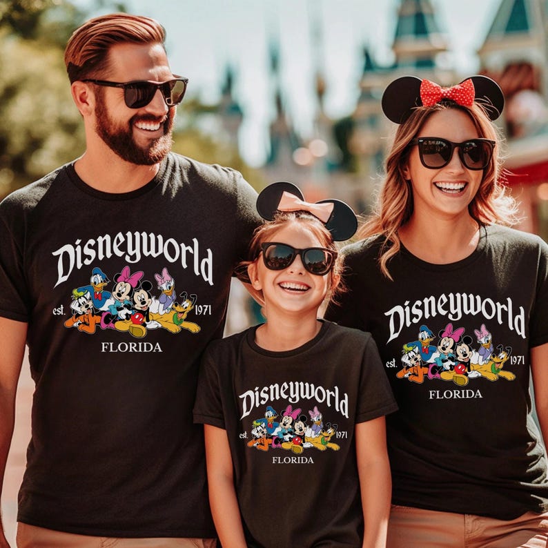 May include: Black short-sleeved t-shirts featuring the white text Disneyworld est. 1971 FLORIDA and a colorful graphic of cartoon characters including Mickey Mouse, Minnie Mouse, Donald Duck, Daisy Duck, and Pluto, worn by three individuals.