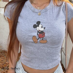 K&ouml;nnte beinhalten: Graues, kurz&auml;rmeliges Crop-Top mit einer Grafik von Mickey Mouse in seinem klassischen Outfit. Das Shirt hat einen Rundhalsausschnitt und ist aus einem weichen, melierten Stoff gefertigt. Der Text "Athletic Heather" ist unten sichtbar.