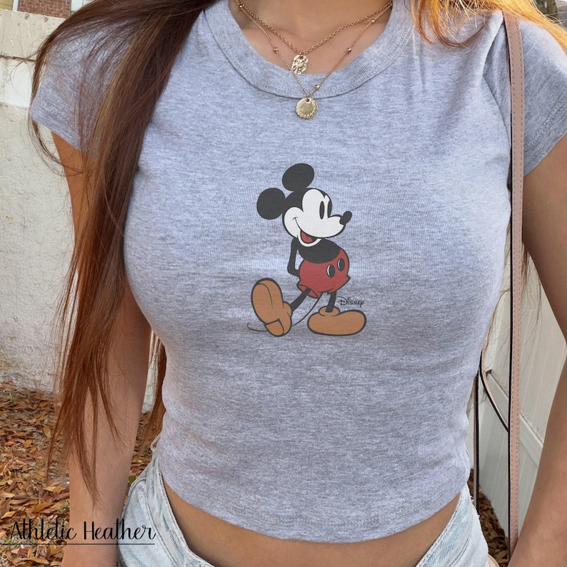 K&ouml;nnte beinhalten: Graues, kurz&auml;rmeliges Crop-Top mit einer Grafik von Mickey Mouse in seinem klassischen Outfit. Das Shirt hat einen Rundhalsausschnitt und ist aus einem weichen, melierten Stoff gefertigt. Der Text "Athletic Heather" ist unten sichtbar.