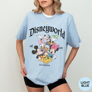 May include: A light blue short-sleeve t-shirt featuring a graphic of Disney characters including Mickey Mouse, Minnie Mouse, Donald Duck, Daisy Duck, Goofy, and Pluto. The text reads Disneyworld, est. 1971, and FLORIDA in black lettering.