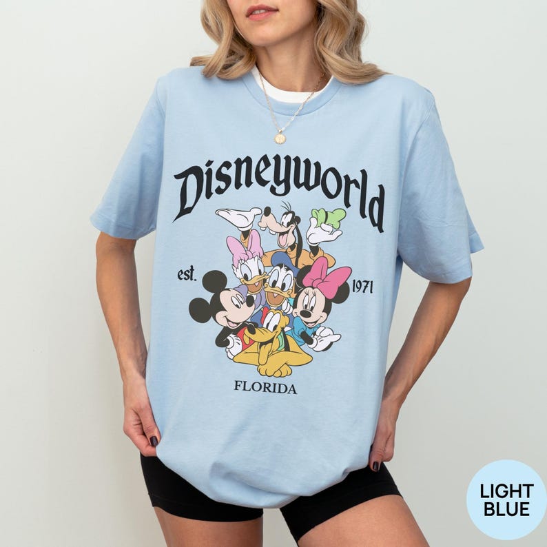 May include: A light blue short-sleeve t-shirt featuring a graphic of Disney characters including Mickey Mouse, Minnie Mouse, Donald Duck, Daisy Duck, Goofy, and Pluto. The text reads Disneyworld, est. 1971, and FLORIDA in black lettering.