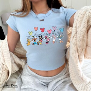 May include: Baby blue crop top featuring a graphic of Disney characters holding Mickey Mouse-shaped balloons. The characters include Pluto, Goofy, Mickey, Minnie, Daisy, and Donald Duck. The text "Baby Blue" is at the bottom of the image.