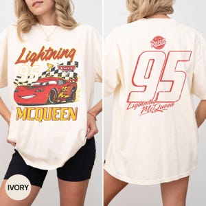 May include: Ivory-colored t-shirt featuring a graphic of Lightning McQueen from the Cars movie. The front design includes the text "Lightning 95 McQueen" with a checkered flag and a red race car. The back displays "95" and "Lightning McQueen".