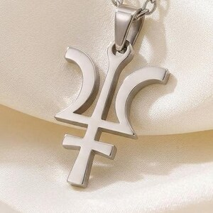 May include: A silver-colored pendant necklace featuring a stylized symbol. The pendant has a unique design with curved and straight lines. The necklace chain is also silver and hangs against a white, silky background. The jewelry is likely made of stainless steel.
