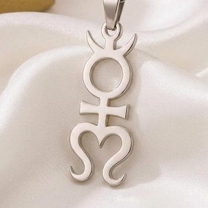 May include: A silver-colored pendant necklace featuring a stylized symbol with a circle, cross, and heart shape. The necklace hangs on a delicate chain, resting on a white satin fabric. A gold and white gift box is in the background.