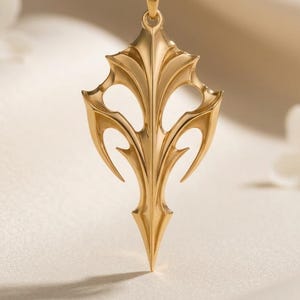 May include: A gold-toned pendant necklace featuring a symmetrical, pointed, shield-like design with intricate cut-out details. The pendant hangs from a delicate gold-toned chain, showcasing a polished metallic finish and sharp, angular edges.