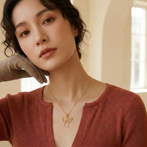 Pode incluir: Um colar dourado com um pingente de anjo. O pingente apresenta um design vazado de um anjo com as asas abertas. O colar &eacute; usado sobre uma blusa de cor ferrugem.