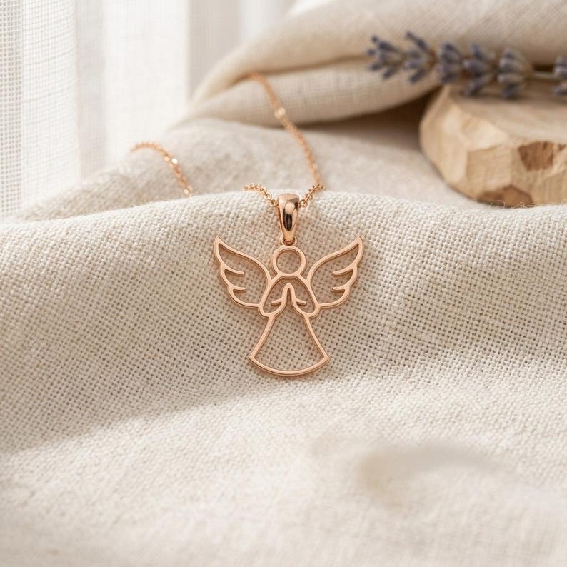 Pode incluir: Um colar com pingente de anjo em ouro rosa. O pingente apresenta um anjo estilizado com asas abertas e m&atilde;os juntas. O colar &eacute; exibido em um tecido de cor neutra, com lavanda ao fundo.