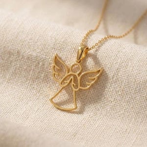 Pode incluir: Um colar com pingente de anjo dourado. O pingente apresenta um design vazado de um anjo com as asas abertas. O colar &eacute; exibido em um tecido texturizado branco-sujo.