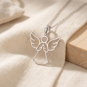 Pode incluir: Um colar com pingente de anjo prateado. O pingente apresenta um design vazado de um anjo com asas abertas e m&atilde;os juntas. A corrente do colar &eacute; delicada e prateada, sobre um tecido bege.