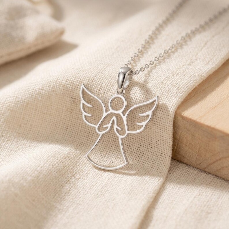 Pode incluir: Um colar com pingente de anjo prateado. O pingente apresenta um design vazado de um anjo com asas abertas e m&atilde;os juntas. A corrente do colar &eacute; delicada e prateada, sobre um tecido bege.