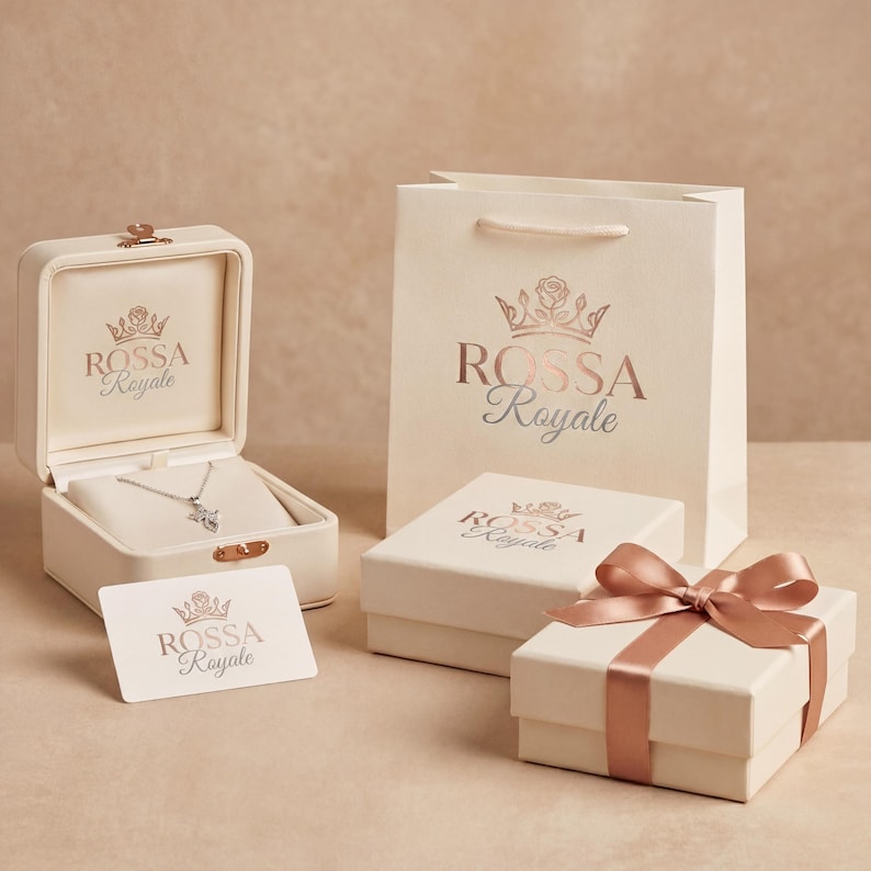 Pode incluir: Uma caixa de joias creme aberta com um colar de prata dentro, uma sacola de presente e duas caixas creme. As caixas e a sacola t&ecirc;m o texto "ROSSA Royale" impresso em ouro rosa. Uma caixa de presente &eacute; amarrada com uma fita de ouro rosa.