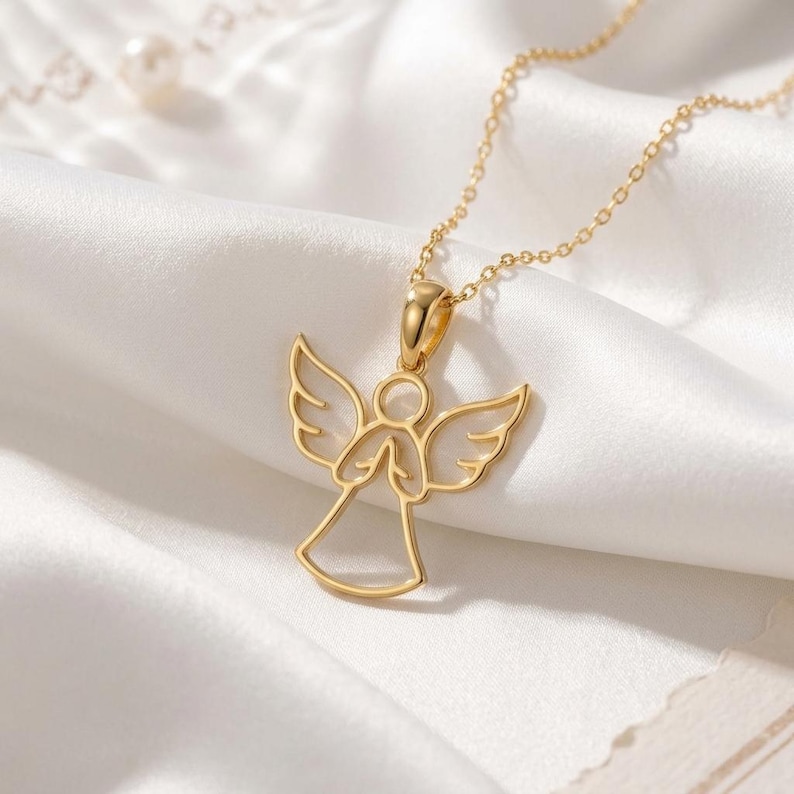 A delicate gold-toned Guardian Angel Necklace featuring a minimalist outline of an angel with outstretched wings and hands clasped in prayer, suspended from a fine cable chain. The sterling silver pendant casts a soft shadow on a light, neutral surface, highlighting its elegant and spiritual design. The image captures the necklace's subtle shine and refined craftsmanship, emphasizing its role as a symbolic piece of minimalist jewelry.
