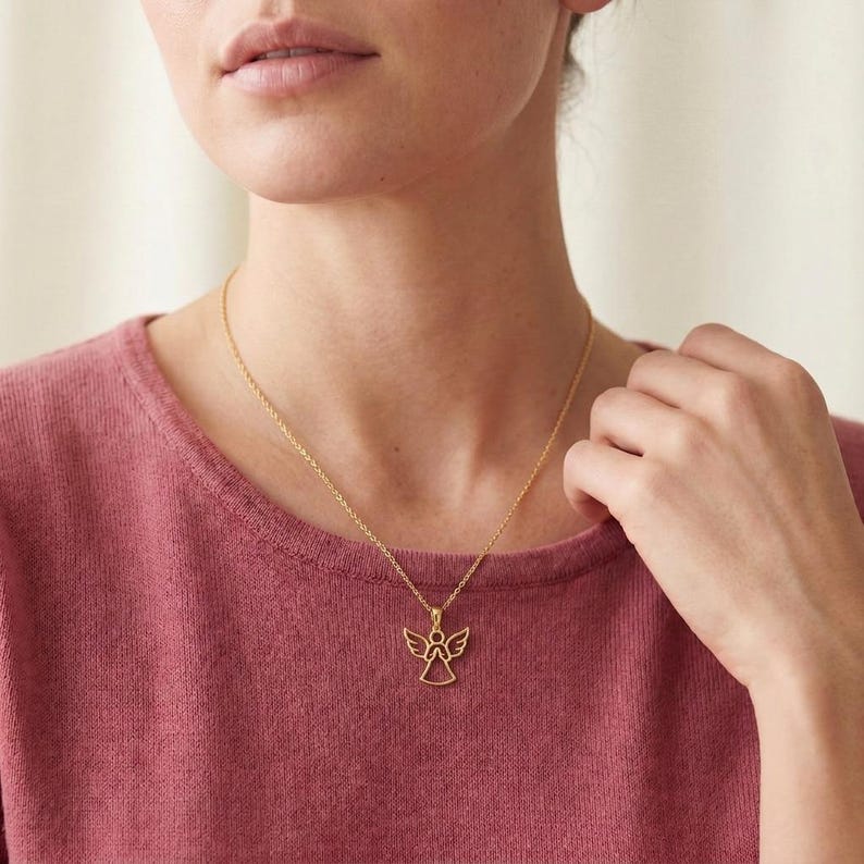 Pode incluir: Um colar dourado com um pingente de anjo. O pingente tem um design aberto com asas e um halo. O colar &eacute; usado por uma pessoa que veste uma camisa rosa.