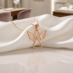 Pode incluir: Um colar com pingente de anjo em ouro rosa. O pingente apresenta um design vazado de um anjo com asas abertas e m&atilde;os juntas. O colar &eacute; exibido em um tecido branco e sedoso.
