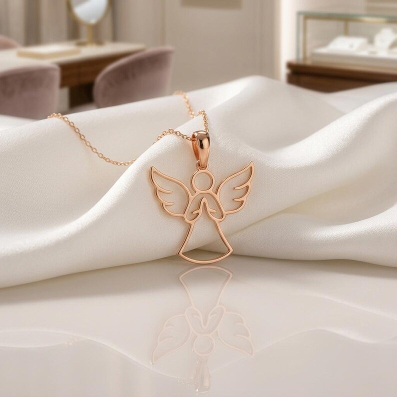 Pode incluir: Um colar com pingente de anjo em ouro rosa. O pingente apresenta um design vazado de um anjo com asas abertas e m&atilde;os juntas. O colar &eacute; exibido em um tecido branco e sedoso.