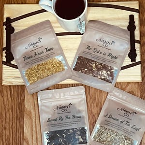 Op de afbeelding: Een houten dienblad met een witte theemok en vier kleine zakjes met theemengsels. De zakjes zijn gelabeld met "Three Hour Tour", "The Spice is Right", "Saved By The Brew" en "I Dream of Tea Leaf".