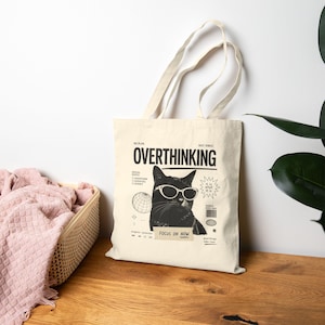 Puede incluir: Bolso tote de color crema con un gato negro con gafas de sol y la palabra "OVERTHINKING" en negrita. El bolso tiene un diseño gráfico con el texto "FOCUS ON NOW" y otras pequeñas ilustraciones.