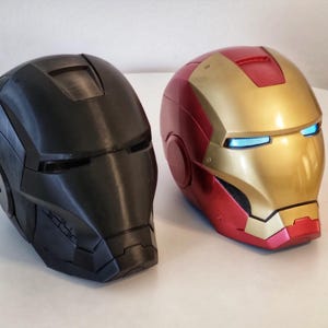 Könnte beinhalten: Zwei Vollvisierhelme, einer in mattem Schwarz und der andere in metallischem Rot und Gold. Der rot-goldene Helm hat leuchtend blaue Augenschlitze, während der schwarze Helm dunkle, nicht beleuchtete Augenschlitze und eine glatte Oberfläche aufweist.