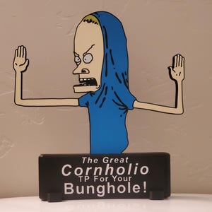 May include: A cartoon cutout of a character with a blue shirt and hood, arms outstretched. The character has a pale face, black eyes, and an open mouth. Below, a black rectangular sign reads "The Great Cornholio TP For Your Bunghole!"