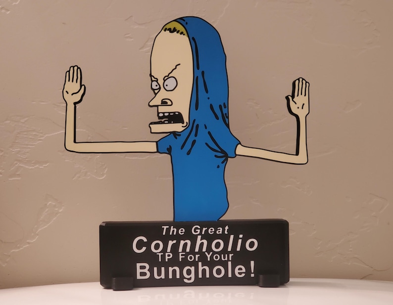 May include: A cartoon cutout of a character with a blue shirt and hood, arms outstretched. The character has a pale face, black eyes, and an open mouth. Below, a black rectangular sign reads "The Great Cornholio TP For Your Bunghole!"