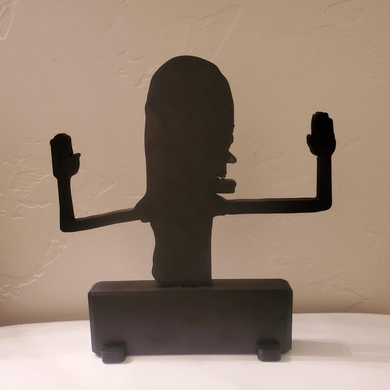 May include: Black metal sculpture of a cartoon character with arms outstretched and hands up. The sculpture has a rectangular base with small feet. The character has a long head and a prominent nose.