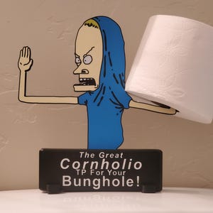 May include: A novelty toilet paper holder featuring a cartoon character in a blue shirt and hood, holding a roll of white toilet paper. The base is a black rectangle with the text "The Great Cornholio TP For Your Bunghole!"