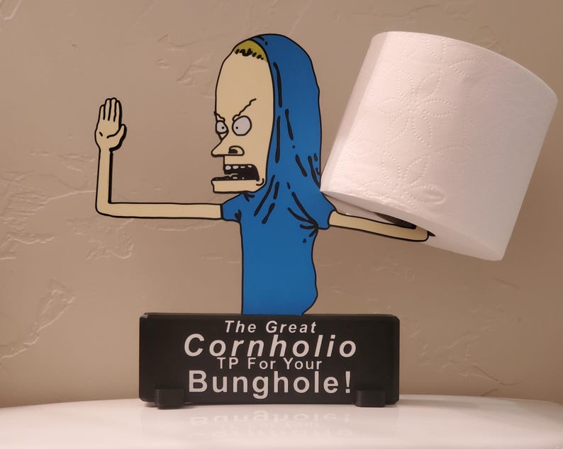 May include: A novelty toilet paper holder featuring a cartoon character in a blue shirt and hood, holding a roll of white toilet paper. The base is a black rectangle with the text "The Great Cornholio TP For Your Bunghole!"