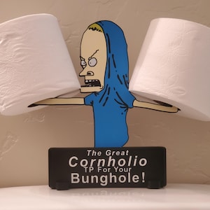 May include: A novelty toilet paper holder featuring a cartoon character in a blue shirt and hood, holding two rolls of white toilet paper. The black base reads "The Great Cornholio TP For Your Bunghole!"